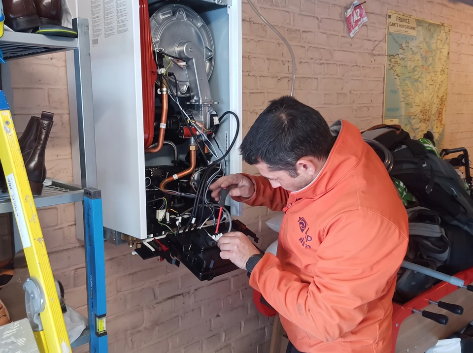 Technicien qualifié en tenue professionnelle complète, avec gilet de travail ou polo bleu foncé marqué du logo de l'entreprise, effectuant un diagnostic ou une maintenance sur une chaudière murale gaz condensation Viessmann dans un local technique résidentiel. Il doit consulter ou vérifier un document (cahier technique, attestation de conformité ou fiche de certification), montrant ainsi l'aspect professionnel et réglementaire de son intervention. Arrière-plan montrant des installations techniques (tuyauterie cuivre/polypropylène, raccordements aux normes, ballon tampon). Éclairage naturel ou artificiel professionnel mettant en avant la compétence et la technicité.