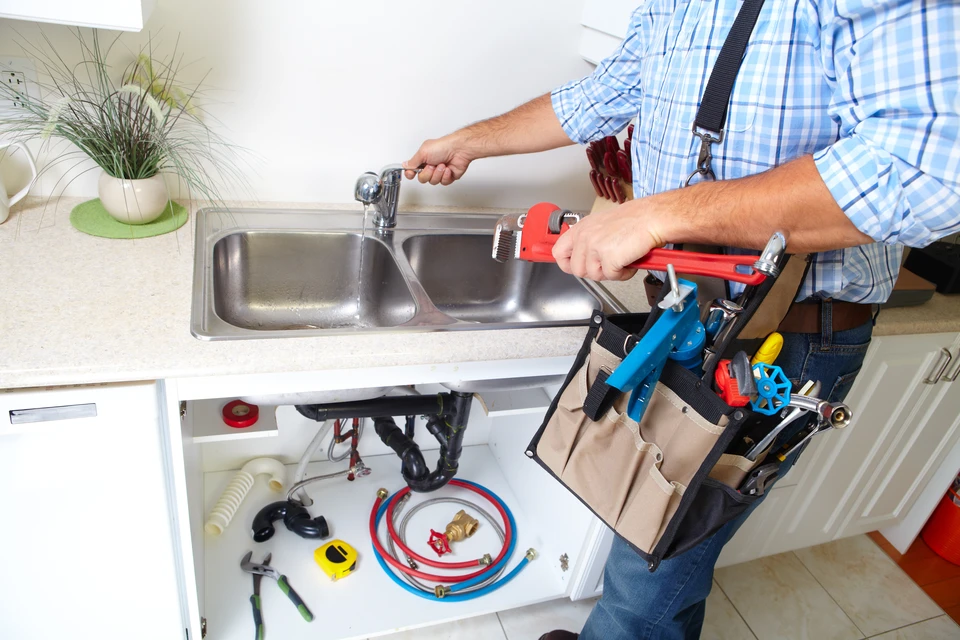 Plombier professionnel intervenant dans une cuisine moderne équipée, tenant des flexibles de raccordement colorés près d'un évier double bac en inox