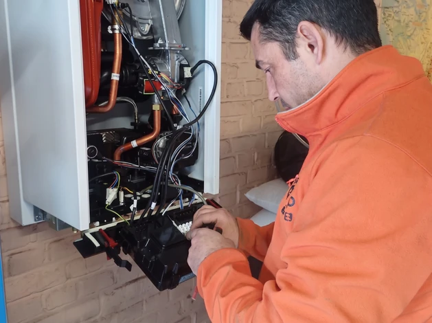 Un artisan expérimenté en tenue de travail professionnelle (gilet ou polo de marque) effectuant une installation de plomberie-sanitaire dans une salle de bain en cours de rénovation. L'artisan est en action, posant ou vérifiant une tuyauterie ou une vasque. L'arrière-plan montre des outils professionnels, du matériel de chantier organisé et des éléments de salle de bain partiellement terminée. L'image capture l'expertise, la proximité et le savoir-faire artisanal dans un contexte professionnel réaliste.