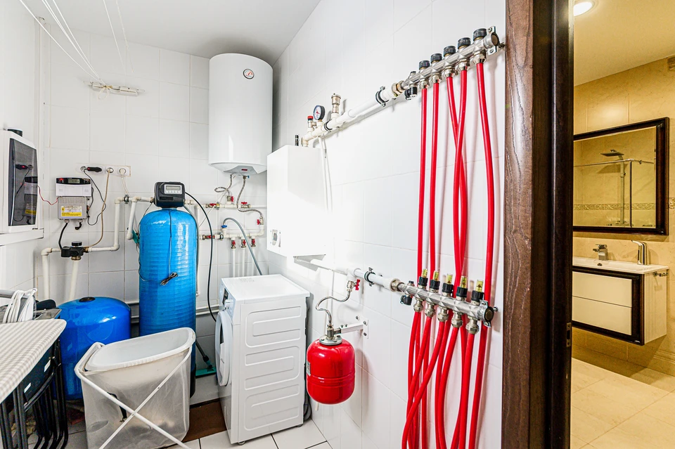 Photographie de chantier d'intérieur montrant une rénovation en cours où plusieurs corps de métier interviennent : au premier plan, des installations électriques visibles (gaine, câblage, boîtiers), au centre un radiateur ou une tuyauterie de chauffage installés, et en arrière-plan des éléments de plomberie (tuyauteries d'eau apparentes ou éléments de douche/baignoire en installation). L'image doit montrer de manière claire et simultanée les trois domaines (électricité, plomberie, chauffage) pour symboliser l'offre intégrée et multi-métier de l'entreprise. La scène doit être réaliste, professionnelle et en cours de réalisation pour mettre en avant la prise en charge globale des travaux.